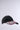 The NAILED IT HAT is a black ball cap showcasing a graphic design of red, white, and gray lipstick tubes on the brim, set against a plain light background.
