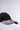 The NAILED IT HAT features a black flat-brim cap adorned with pink, white, and red press-on nail decorations. It is shown against a simple light gray background.