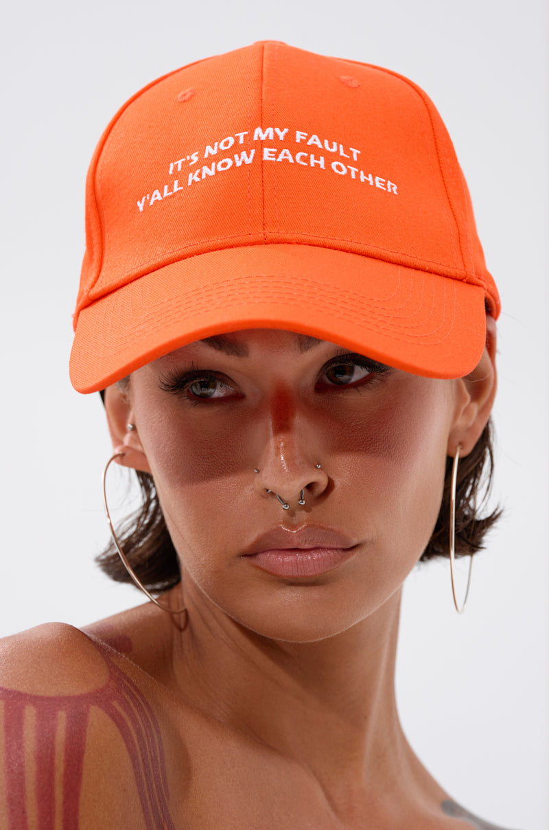 Someone wearing the ITS NOT HAT, a bright orange cotton cap with "IT'S NOT MY FAULT Y'ALL KNOW EACH OTHER" embroidered on it, along with large hoop earrings, facial piercings, and red body paint on their shoulder and nose.