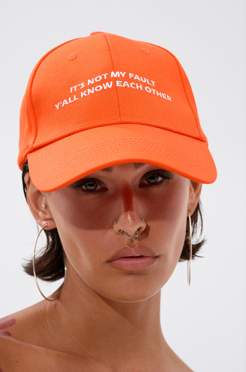 A person with large hoop earrings and facial piercings wears the ITS NOT HAT, an orange cotton cap with an adjustable back strap and “IT'S NOT MY FAULT Y'ALL KNOW EACH OTHER” in white text. The background is plain and light colored.