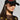 A person wears the WITH LOVE PATCH SNAPBACK, a black hat adorned with red and gold heart designs, paired with large hoop earrings. They have on a sheer black top enhanced with gold embellishments. The plain background highlights the fashion details.