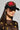 A person wears the WITH LOVE PATCH SNAPBACK, a black hat adorned with red and gold heart designs, paired with large hoop earrings. They have on a sheer black top enhanced with gold embellishments. The plain background highlights the fashion details.