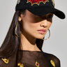 A person wears the WITH LOVE PATCH SNAPBACK, a black hat adorned with red and gold heart designs, paired with large hoop earrings. They have on a sheer black top enhanced with gold embellishments. The plain background highlights the fashion details.