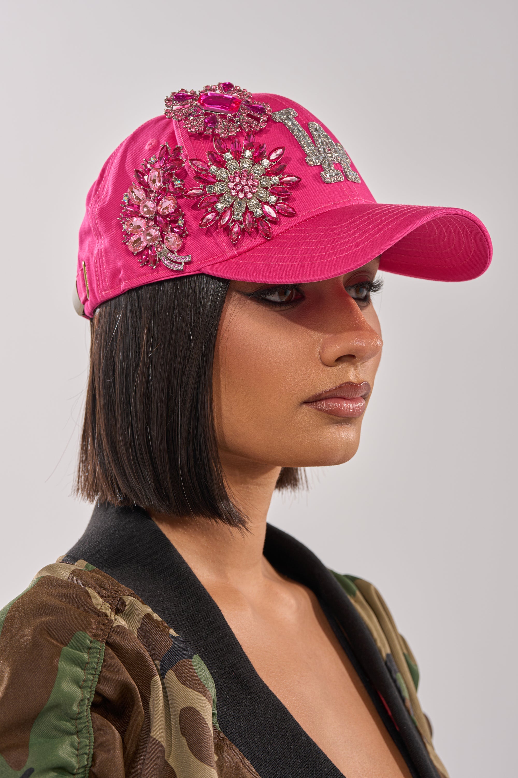 A woman with a bob haircut and nose ring wears the vibrant VAL BROOCH HAT, a hot pink baseball cap detailed with crystal floral appliqués and a sequin letter, paired with a camouflage jacket with a black collar. 0
