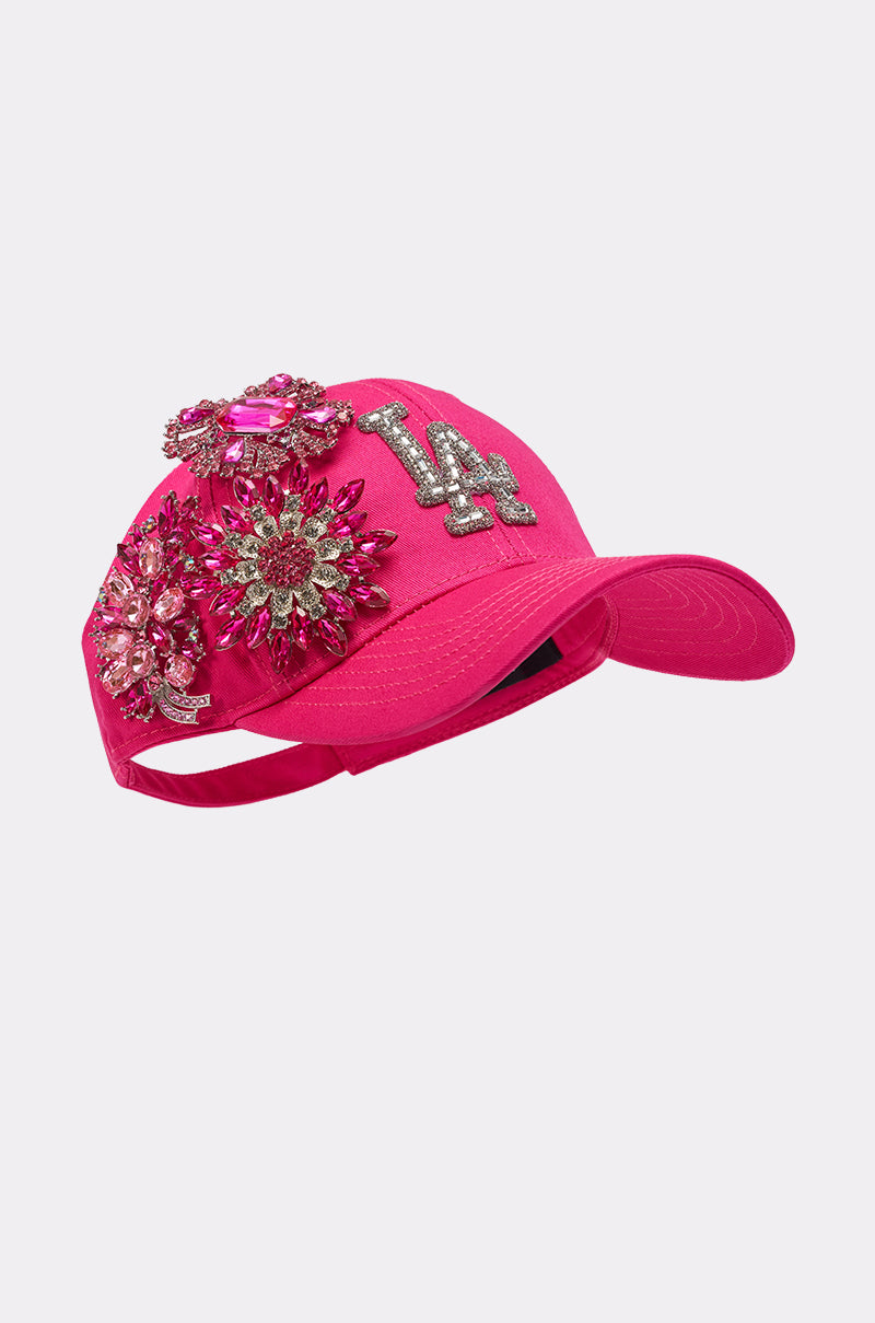 The VAL BROOCH HAT is a hot pink cap adorned with crystal floral appliqués and a sparkling silver "LA" logo, making it a standout rhinestone LA hat with bold jeweled embellishments. 2