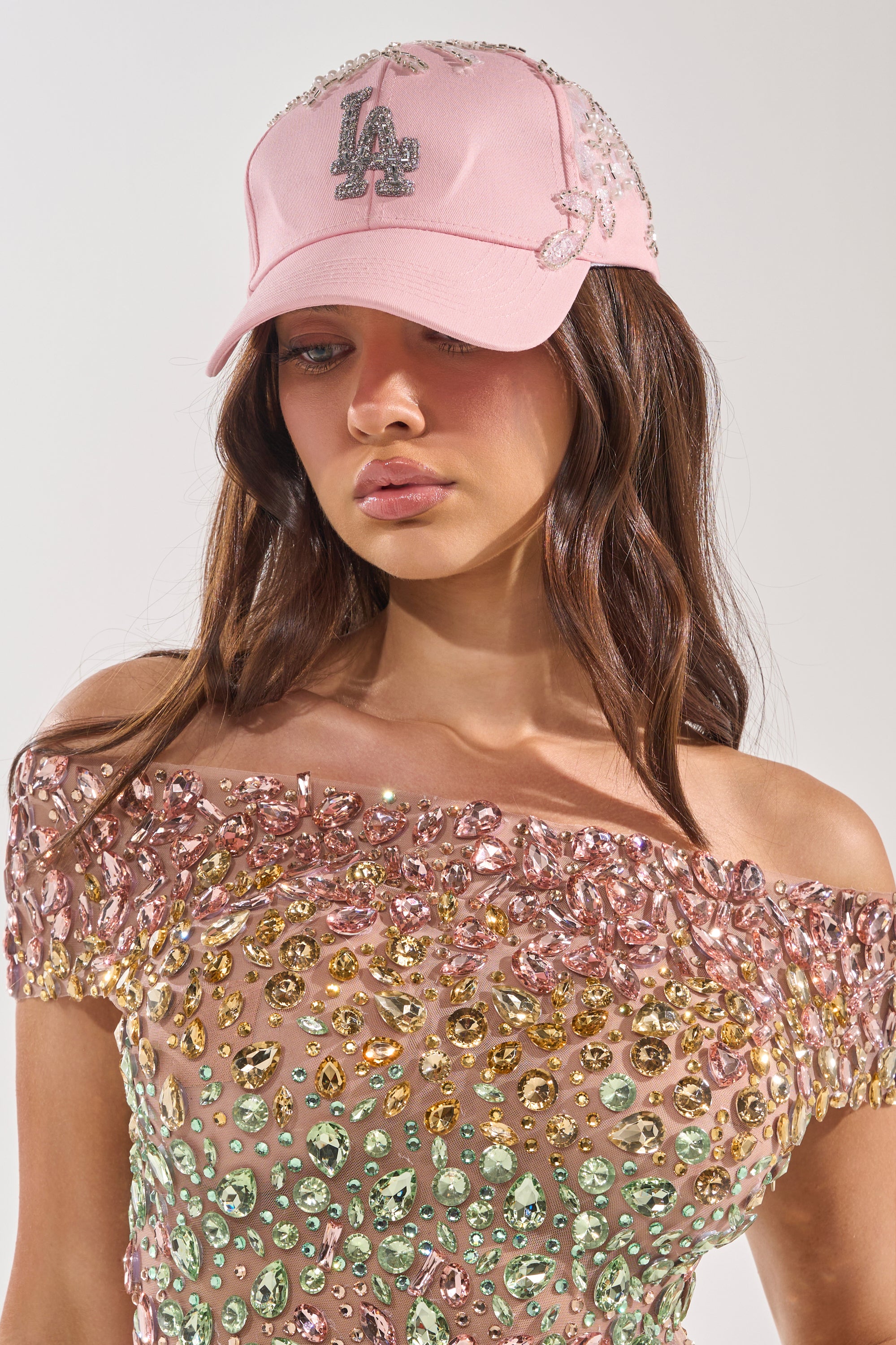 A woman wears THE CITY APPLIQUE HAT in pink, featuring rhinestone embellishments, paired with an off-the-shoulder pink jeweled top. She has long brown hair and a neutral expression. 0