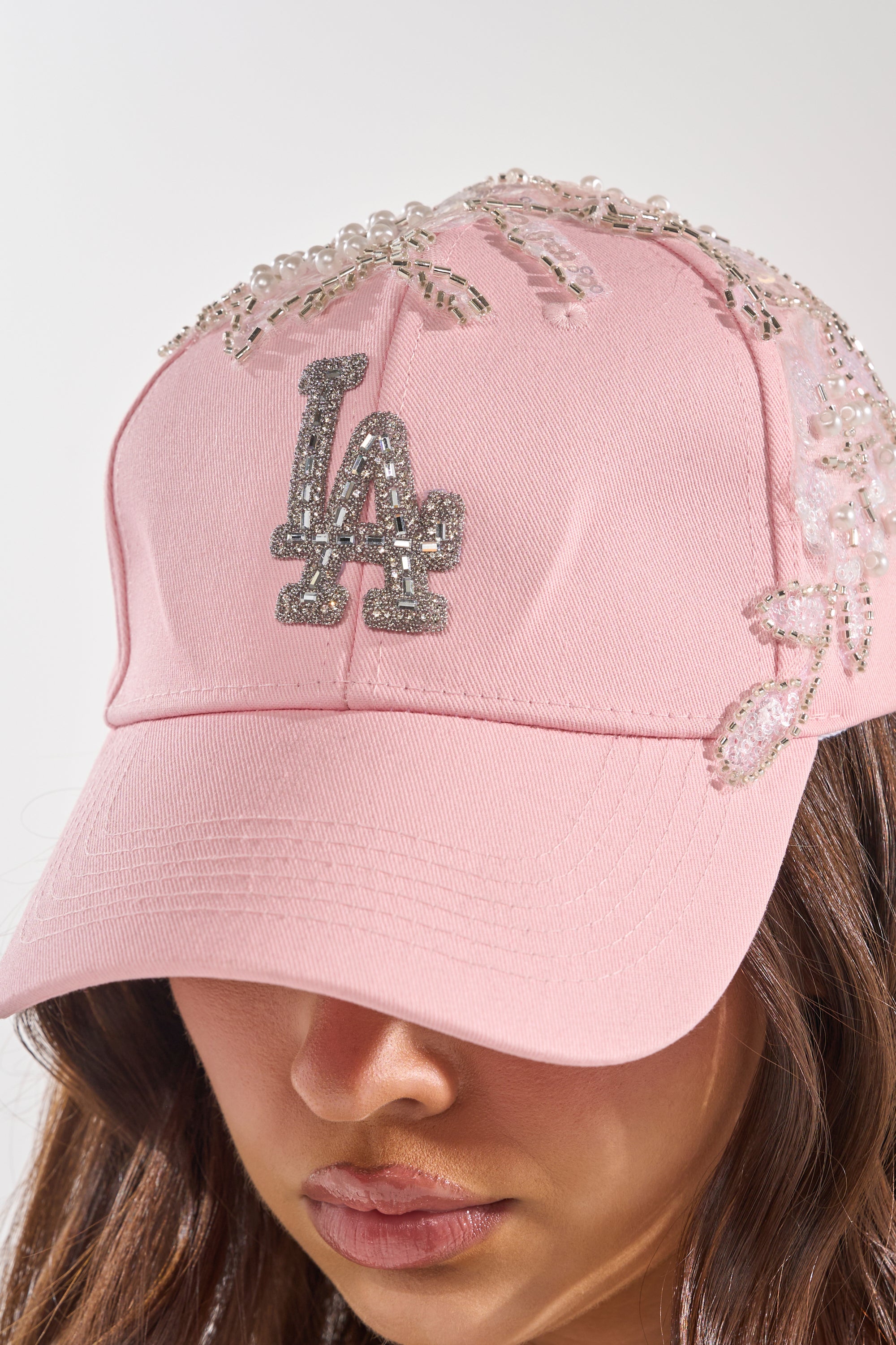 A person wears THE CITY APPLIQUE HAT—a light pink cap with beaded floral accents and a sparkling "LA" logo. Only the lower part of their face is visible. 1