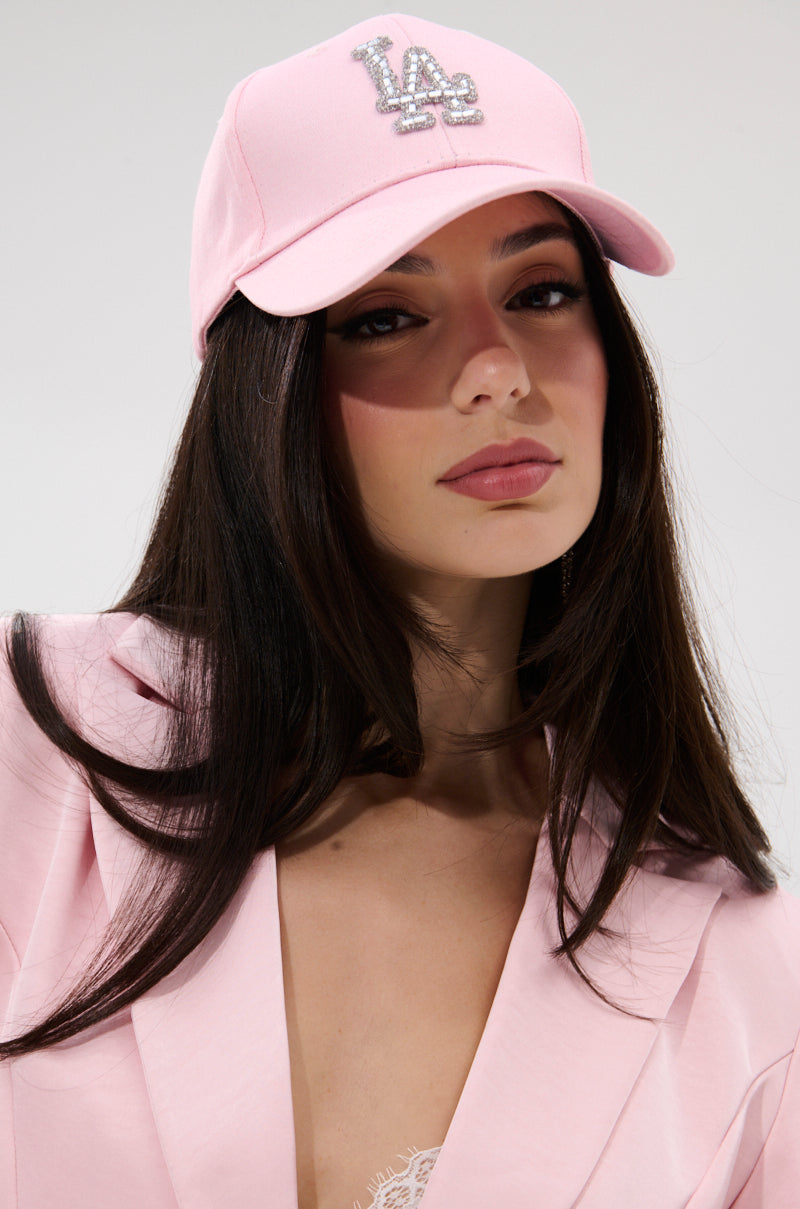 A woman with long dark hair wears the light pink CATCH FLIGHTS HAT and a matching blazer over a lace top, posing against a plain white background.