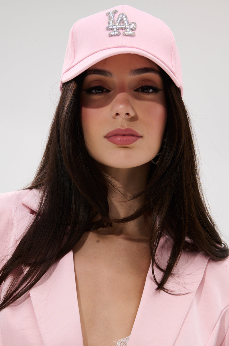 A woman with long dark hair wears a light pink blazer, a lace top, and the CATCH FLIGHTS HAT with a silver “LA” logo. She looks directly at the camera against a plain white background.