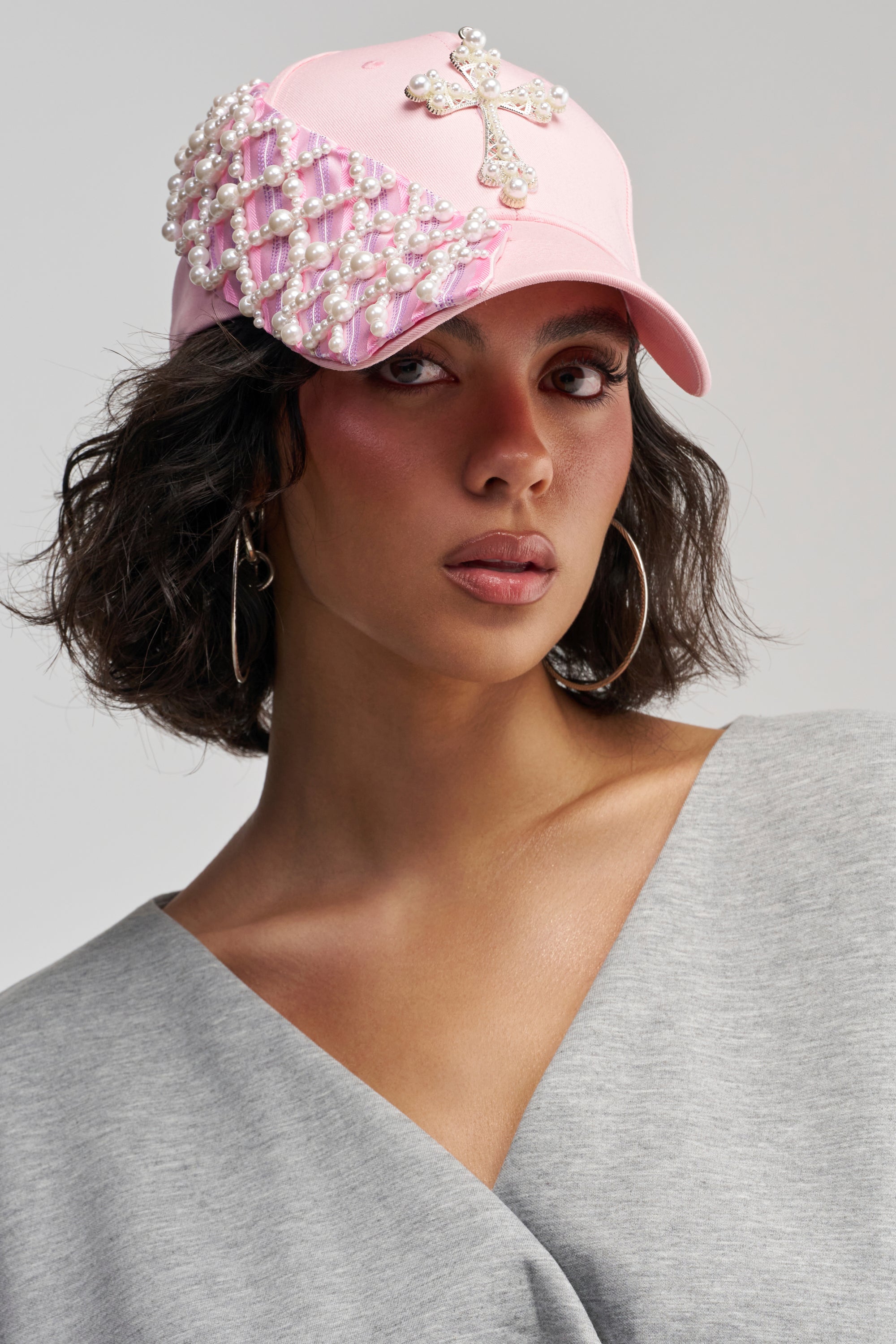 A woman models the TIE IT UP EMBELLISHED BASEBALL HAT in pink, featuring pearl accents, a ribbon detail, and a jeweled cross. She pairs the look with hoop earrings, short dark hair, and a gray V-neck top.