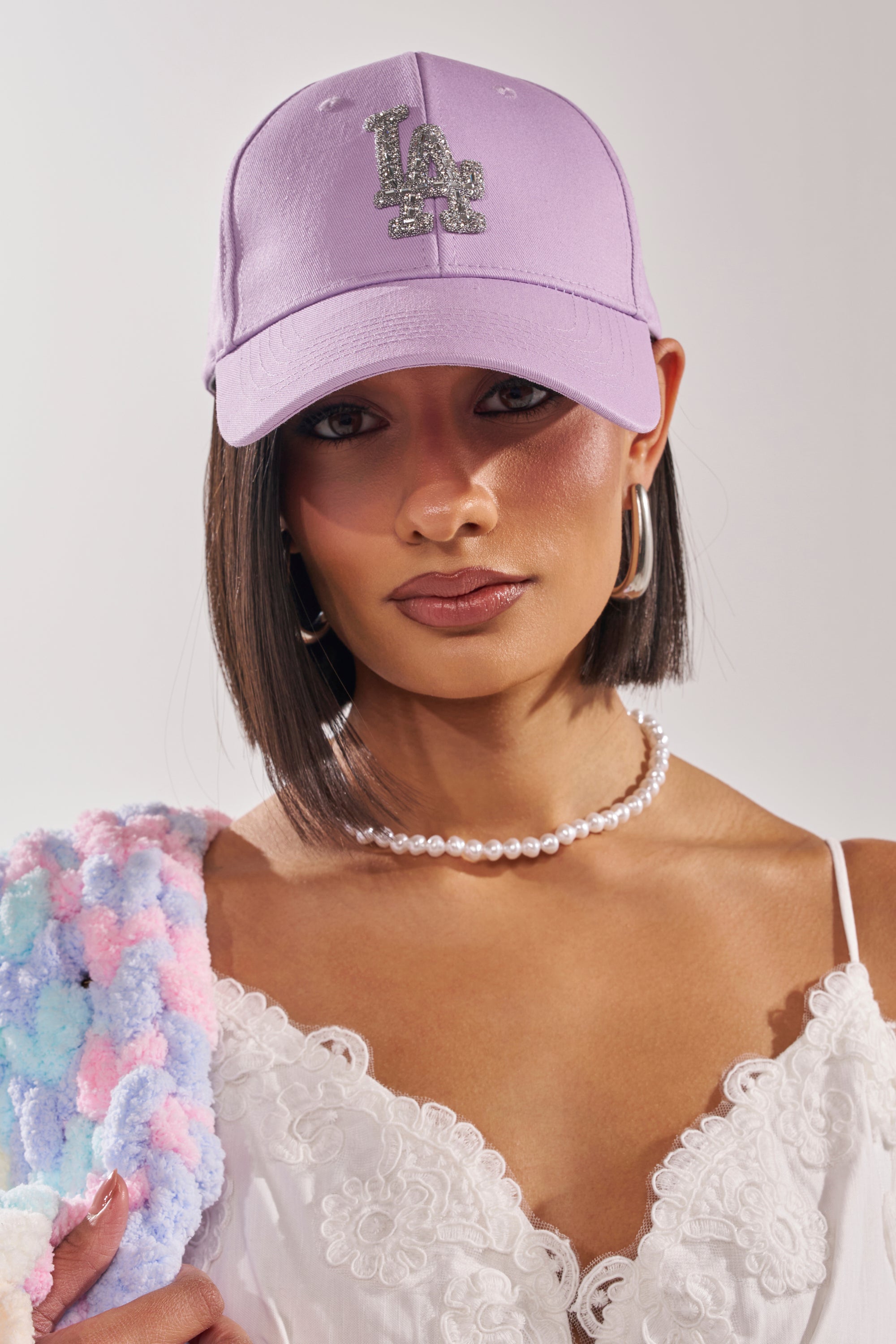 A woman wears the IN THE CITY APPLIQUE HAT, large hoop earrings, a pearl necklace, and a white lace camisole while holding a pastel textured item over her shoulder against a plain background. 0