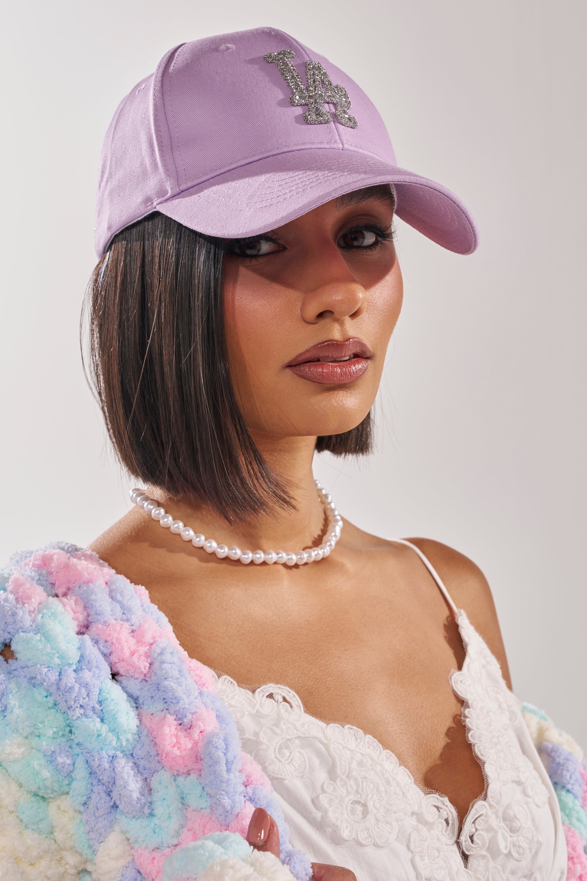 A woman wearing the IN THE CITY APPLIQUE HAT in lavender, a pearl necklace, white lace camisole, and pastel textured jacket poses against a plain light background. 1