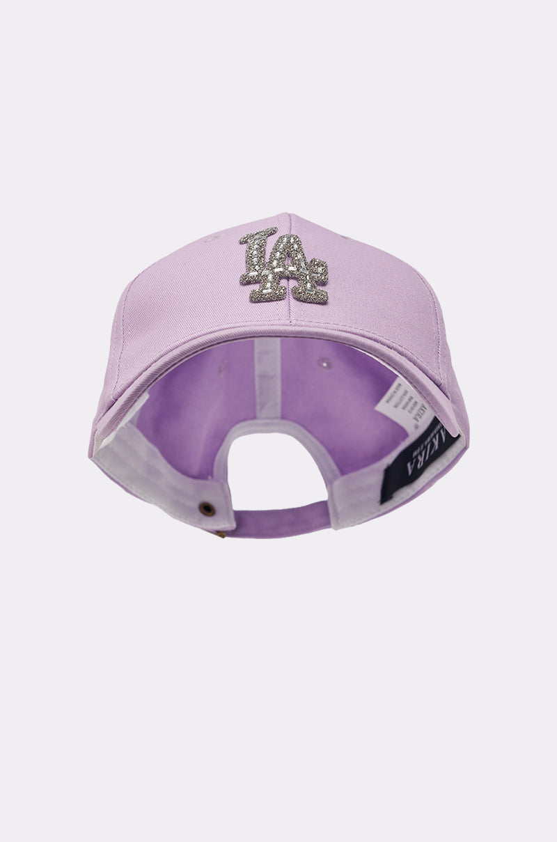 The IN THE CITY APPLIQUE HAT is a lavender baseball cap featuring a glittery rhinestone LA logo on the front, shown from the back with an adjustable strap and visible inside tags. 2