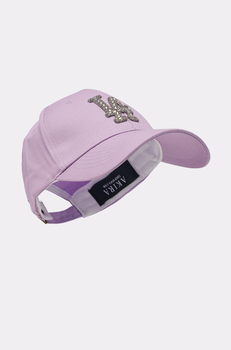 The IN THE CITY APPLIQUE HAT is a lavender baseball cap featuring a silver beaded "LA" crystal logo on the front and an adjustable strap at the back, shown against a plain white background. 3