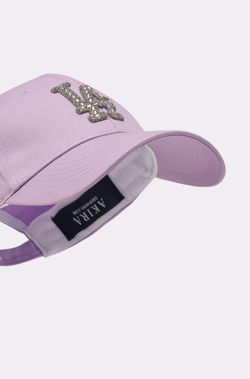 The IN THE CITY APPLIQUE HAT is a lavender baseball cap featuring a glittery crystal "LA" logo on the front and a black "AKIRA" label inside, photographed against a plain light background. 5