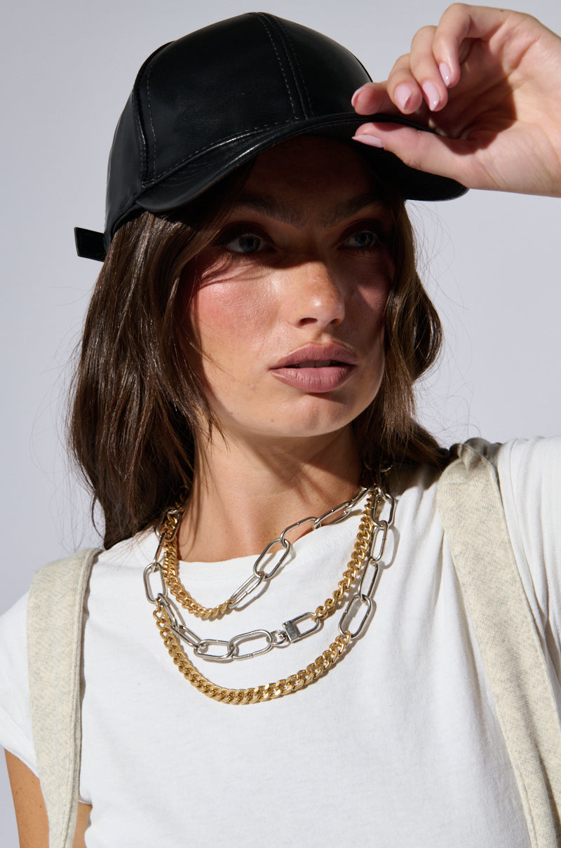 A person with long brown hair, donning a PU SNAPBACK, white t-shirt, and layered gold and silver chains. They are gripping the adjustable strap of the cap with their right hand and gazing to the side with a serious expression set against a neutral background. 0