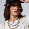 A person with long brown hair, donning a PU SNAPBACK, white t-shirt, and layered gold and silver chains. They are gripping the adjustable strap of the cap with their right hand and gazing to the side with a serious expression set against a neutral background.