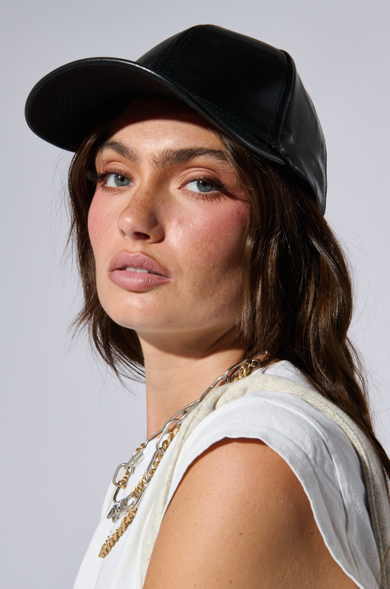 A person with long brown hair wearing a PU SNAPBACK and a white sleeveless top, accessorized with layered gold chains, gazes confidently at the camera. The background is a plain light gray. 1