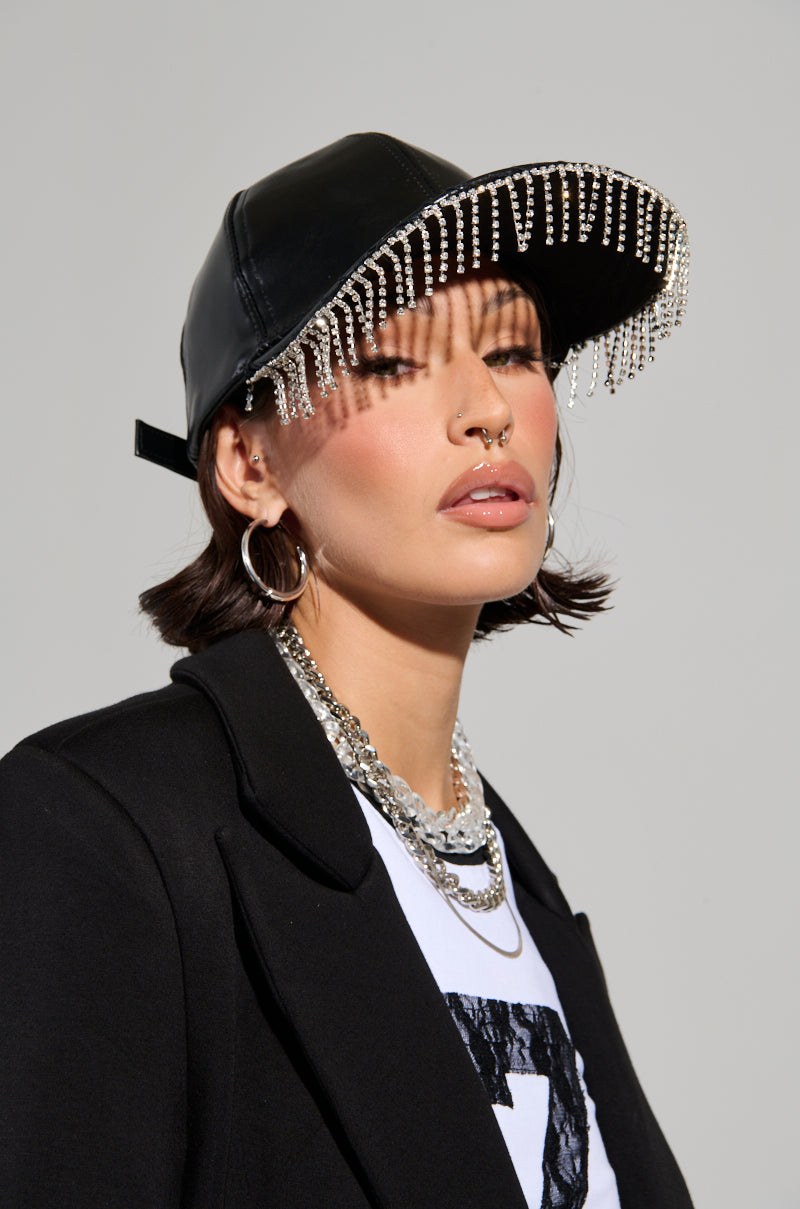 A person wearing the stylish RAINING DIAMONDS FRINGE HAT, a black blazer over a white shirt, and silver jewelry stands against a plain gray background. The ensemble highlights an edgy fashion look with just the right touch of bling.