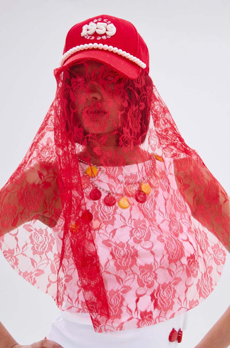 A person wears the ROSE CITY VEIL HAT from AKIRA Label, featuring red lace and pearl accents, paired with a white top with red and yellow buttons against a plain light background.
