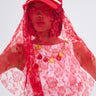 A person wears the ROSE CITY VEIL HAT from AKIRA Label, featuring red lace and pearl accents, paired with a white top with red and yellow buttons against a plain light background.