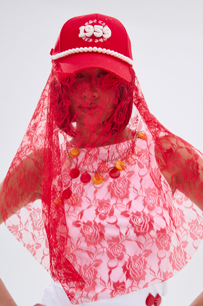Wearing the ROSE CITY VEIL HAT—red lace over a red cap with pearl accents—a person stands against a plain background. A white top with red lace overlay and a red and orange bead necklace complete their look.