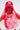 Wearing the ROSE CITY VEIL HAT—red lace over a red cap with pearl accents—a person stands against a plain background. A white top with red lace overlay and a red and orange bead necklace complete their look.