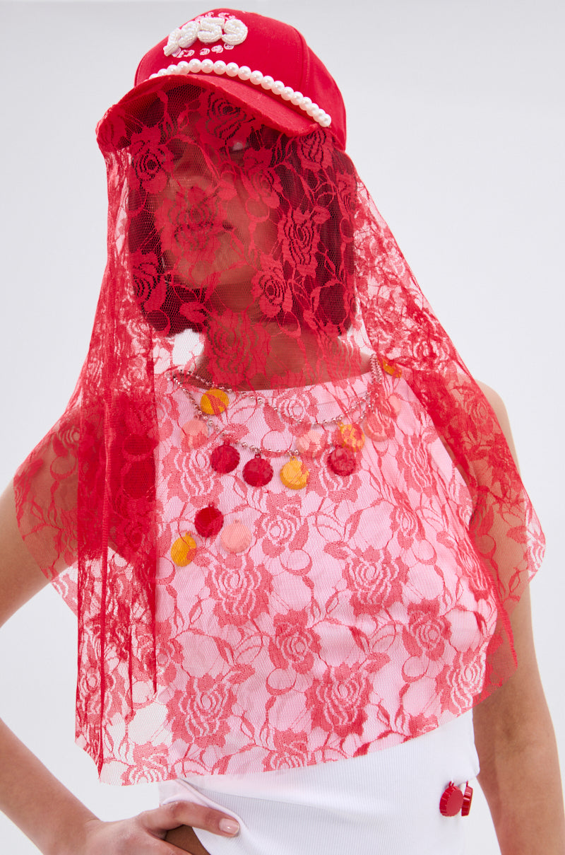 A person wears the ROSE CITY VEIL HAT with pearl embellishments and a red lace veil over their face, paired with a white top featuring red floral lace and colorful buttons, standing against a plain light background.
