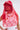 A person wears the ROSE CITY VEIL HAT with pearl embellishments and a red lace veil over their face, paired with a white top featuring red floral lace and colorful buttons, standing against a plain light background.