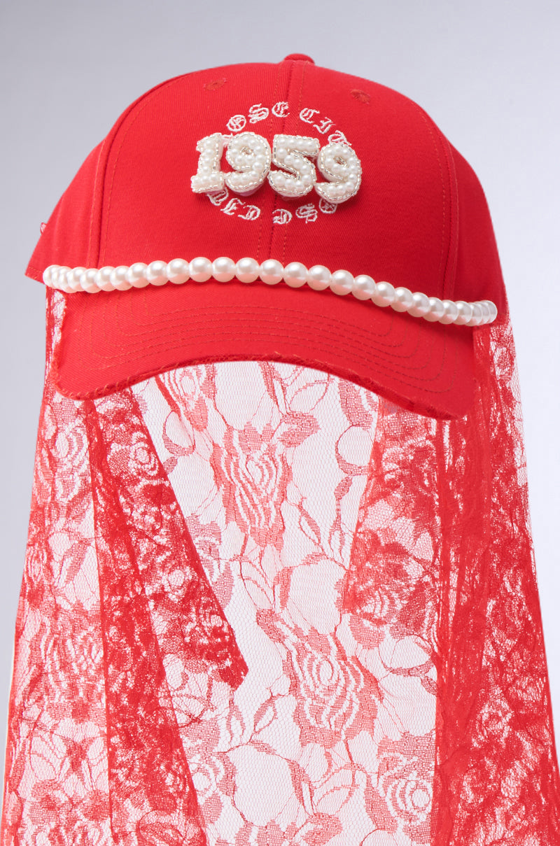 The ROSE CITY VEIL HAT by AKIRA Label is red with "1999" in white textured numbers, pearl accents above the brim, and a red floral lace veil that drapes gracefully.