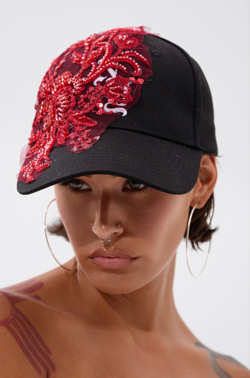 Someone with hoop earrings, facial piercings, and a visible tattoo wears the IN MY CITY APPLIQUE HAT, an adjustable black cotton baseball cap featuring bold floral applique and a large red sequin design on front and side.
