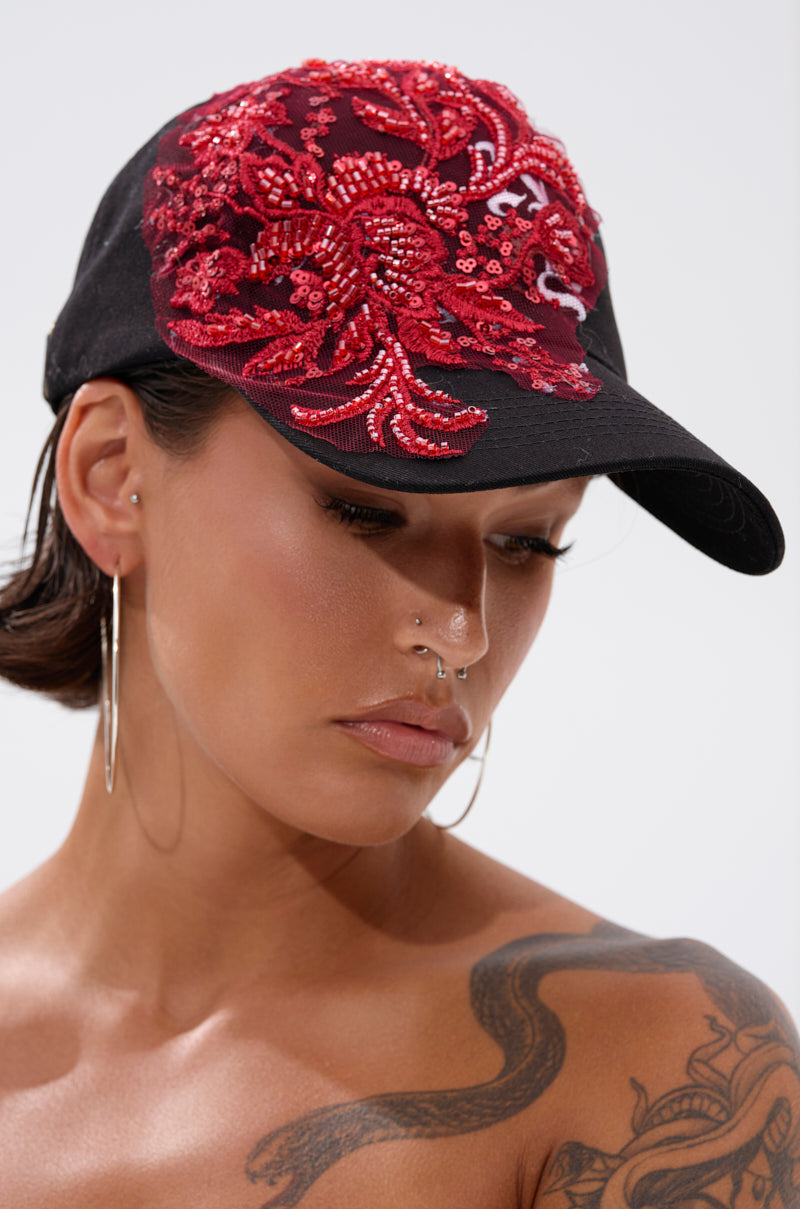 A woman wearing the IN MY CITY APPLIQUE HAT, featuring red sequin and bead floral embroidery, along with large hoop earrings, nose piercings, and a shoulder tattoo, looks downward with a neutral expression.