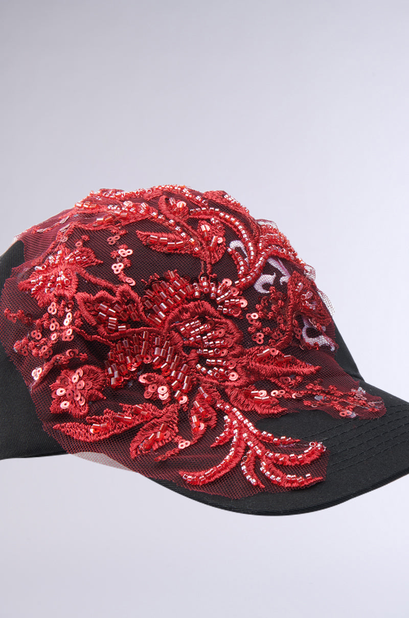 The IN MY CITY APPLIQUE HAT is a black baseball cap adorned with intricate red sequin and bead floral embroidery, showcased against a plain light gray background.