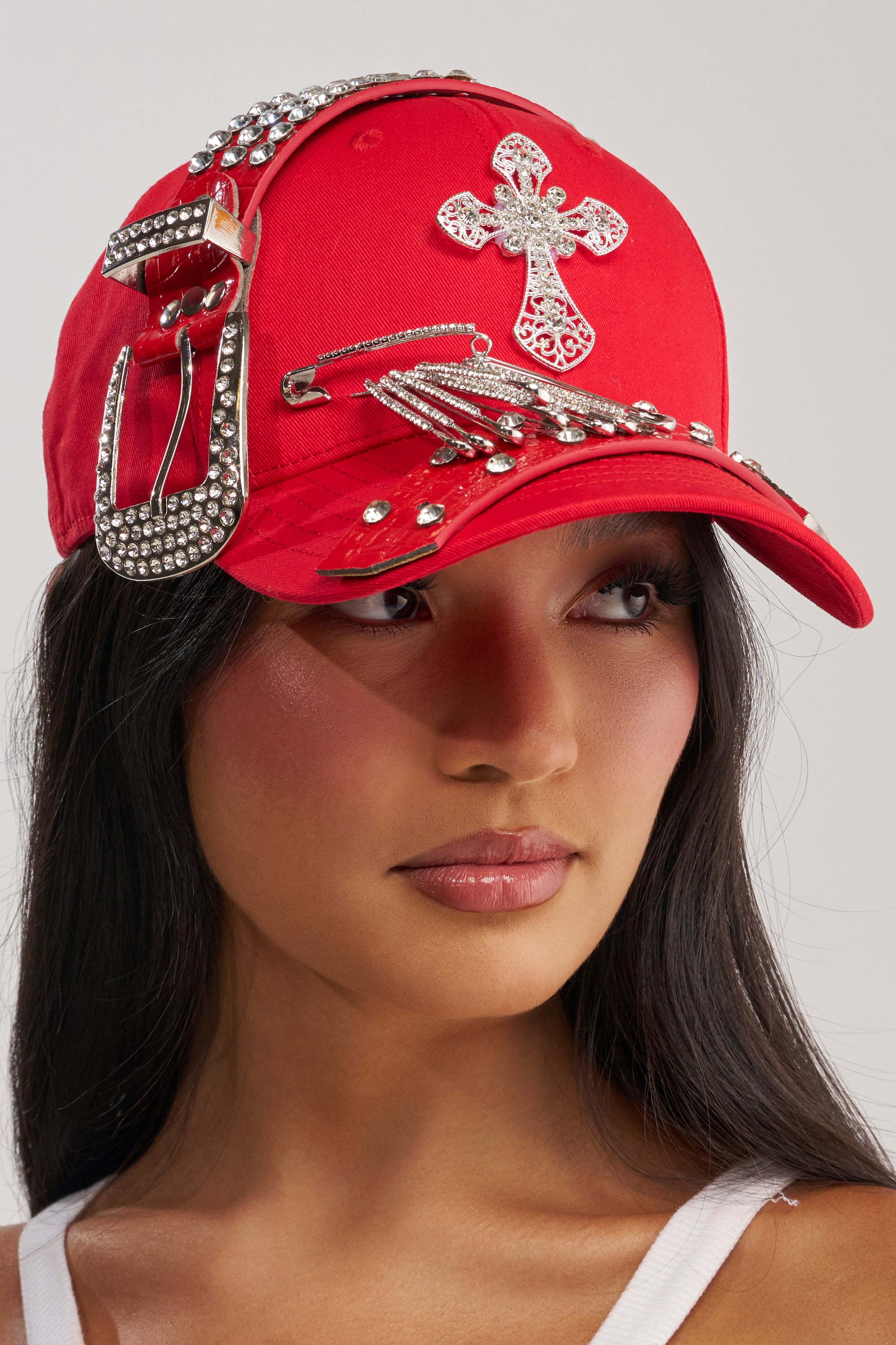 A woman models the BUCKLE UP EMBELLISHED BASEBALL HAT, featuring silver rhinestones, a jeweled cross, a large decorative buckle, and metallic accents, as she gazes to the side against a neutral background.