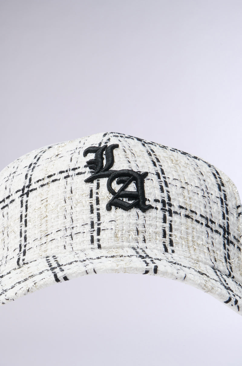 Close-up of the COME TO ME HAT: a white textured cap with a black plaid pattern and bold LA embroidery on the front. This AKIRA Label piece is crafted from tweed fabric and set against a plain, light-colored background. 3