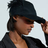 A person in a pinstriped blazer adjusts the brim of their BRB FLYING AWAY HAT, featuring an adjustable back strap and side feather details, against a plain, light background.