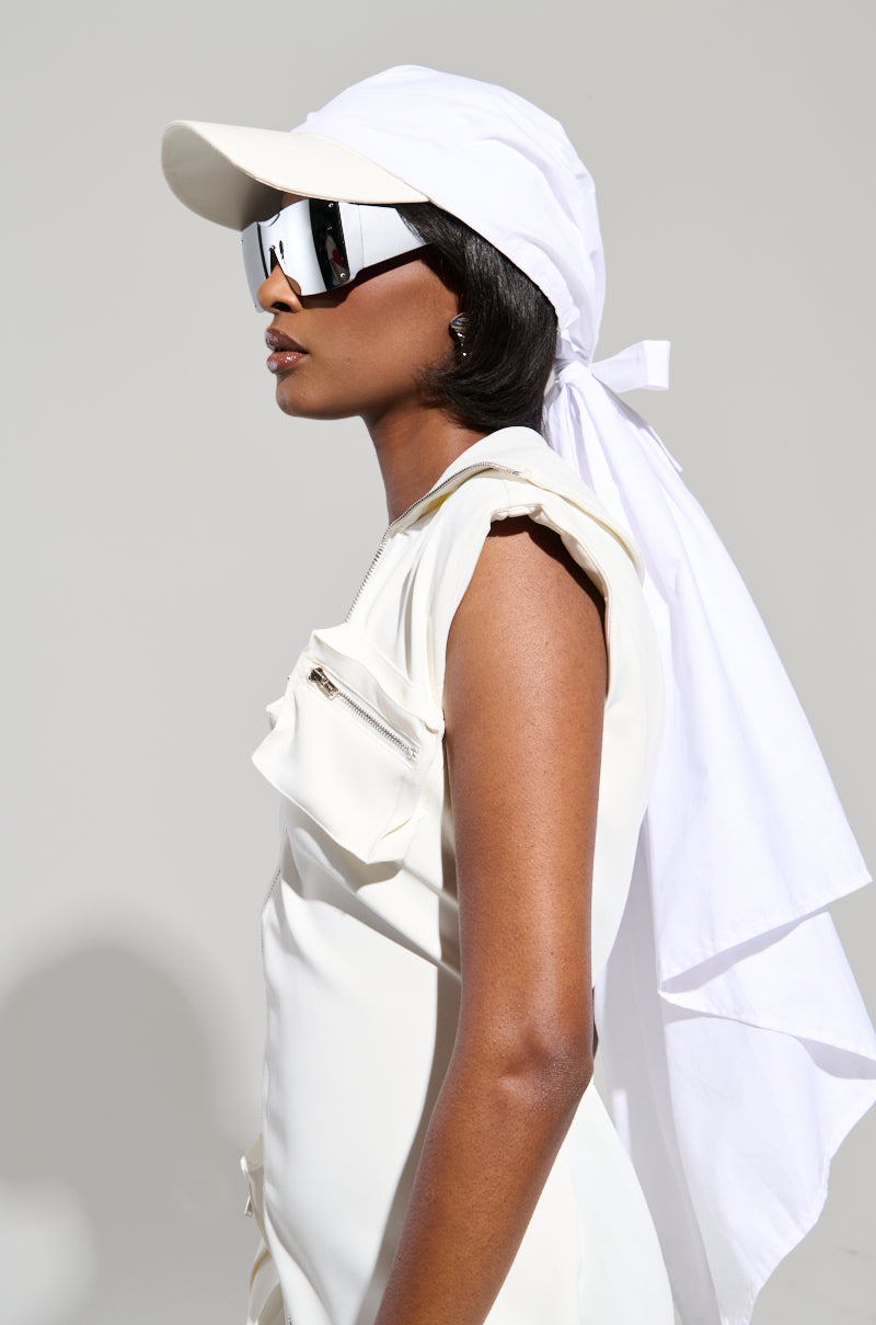 A person in profile is dressed in an AKIRA Label white outfit and is wearing the TIE IT ALL BACK PU HAT, featuring a sleek faux leather design with a long back flap. They complete their look with large, reflective sunglasses against a plain light gray background.