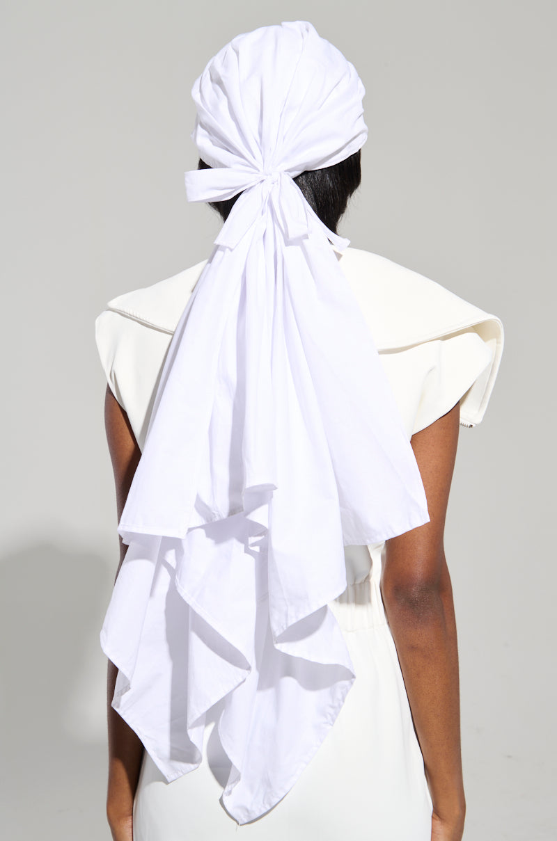 A person wearing the TIE IT ALL BACK PU HAT, featuring a white head wrap with long, flowing fabric cascading down their back, dressed in a striking AKIRA Label outfit with broad shoulders, stands facing away from the camera against a plain background.