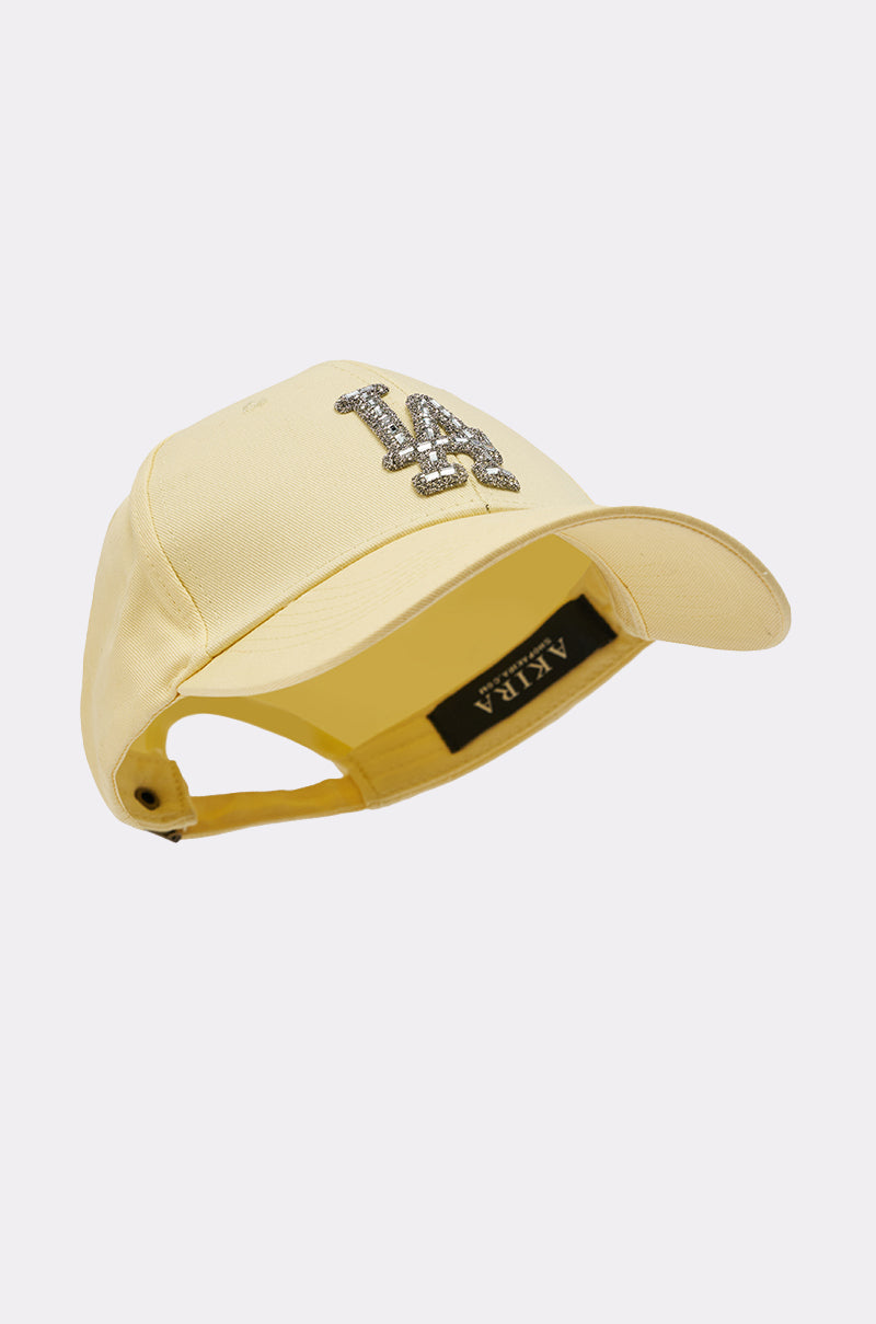 The IN THE SUN BLING HAT is a light yellow adjustable baseball cap with a curved brim, a jeweled "LA" logo on the front, and a black ALKLA label inside. Shown on a white background. 5