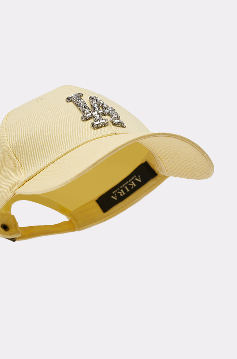The IN THE SUN BLING HAT is a yellow streetwear cap with a curved brim, silver glitter "LA" letters on the front, and a black AKIRA label inside. Displayed sideways on a plain white background. 7