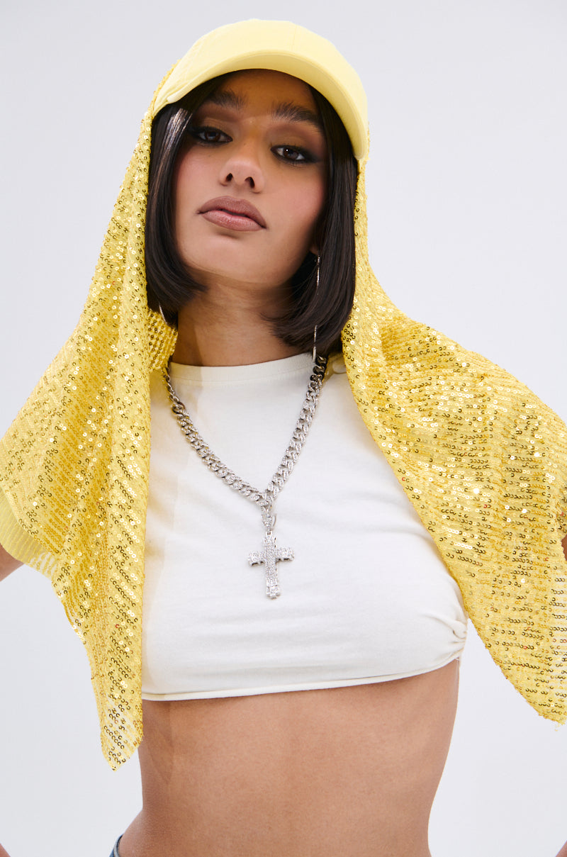 Wearing the IN NYC SEQUIN SCARF HAT draped over a yellow cap, a white crop top, and a chunky silver cross necklace, this person poses confidently against a plain background.