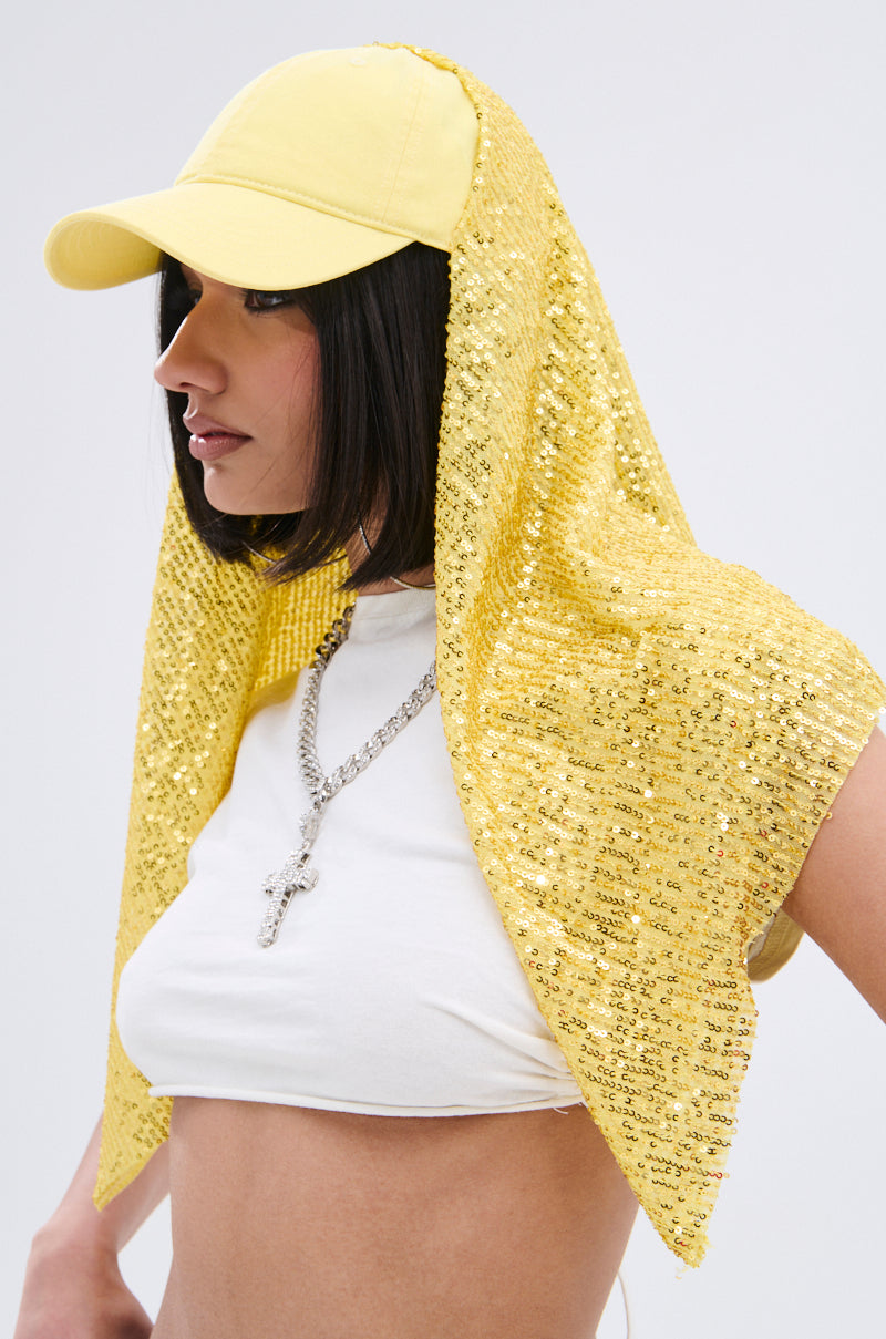 A woman wears the IN NYC SEQUIN SCARF HAT, a yellow scarf hat with tonal sequin embellishments, styled over her head with a white crop top and silver cross necklace, posing against a plain light background.
