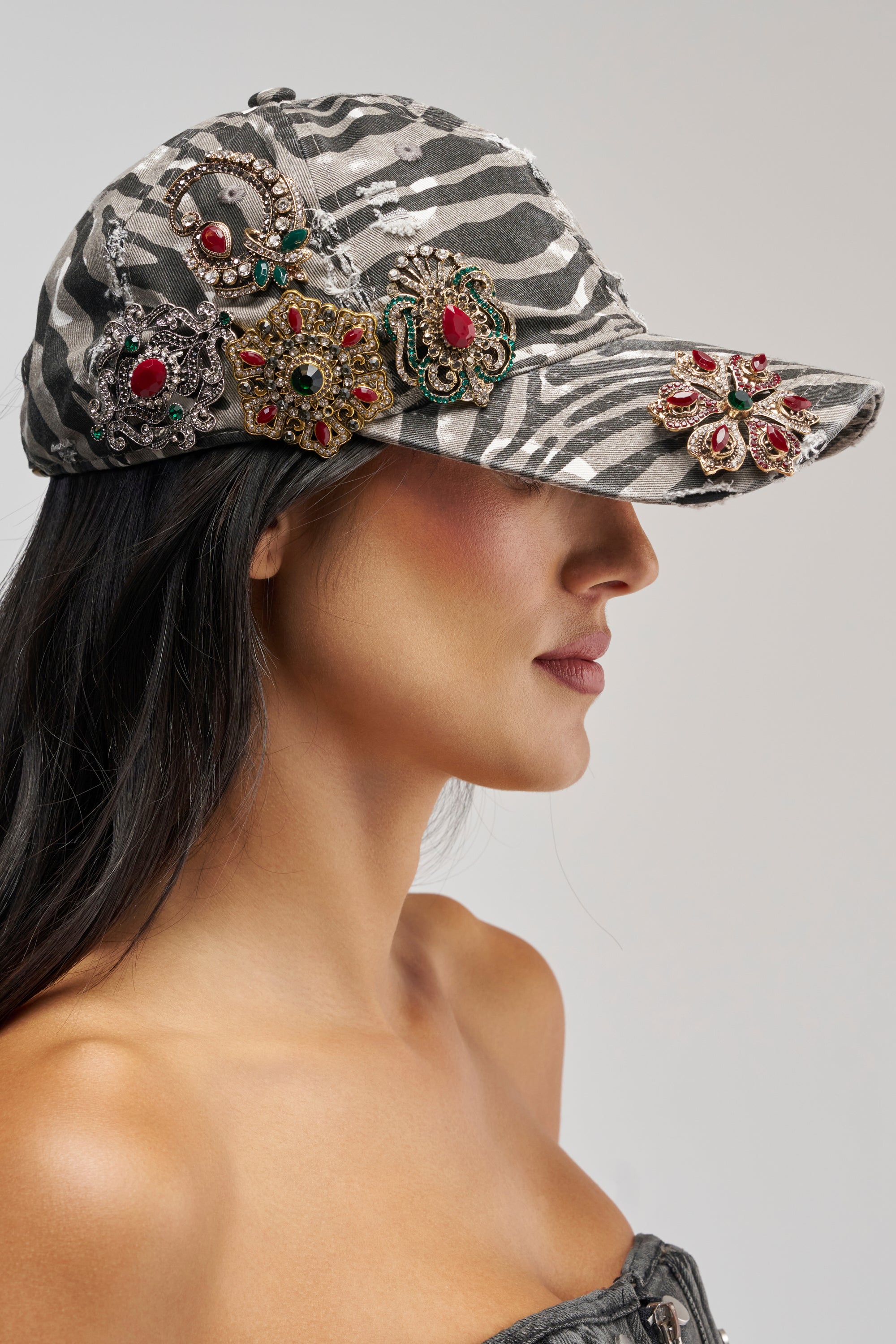 A woman in profile wearing the ZEBRA HAT, a gray and white zebra-striped cap adorned with large, ornate jeweled brooches, paired with a strapless top, set against a light gray background.