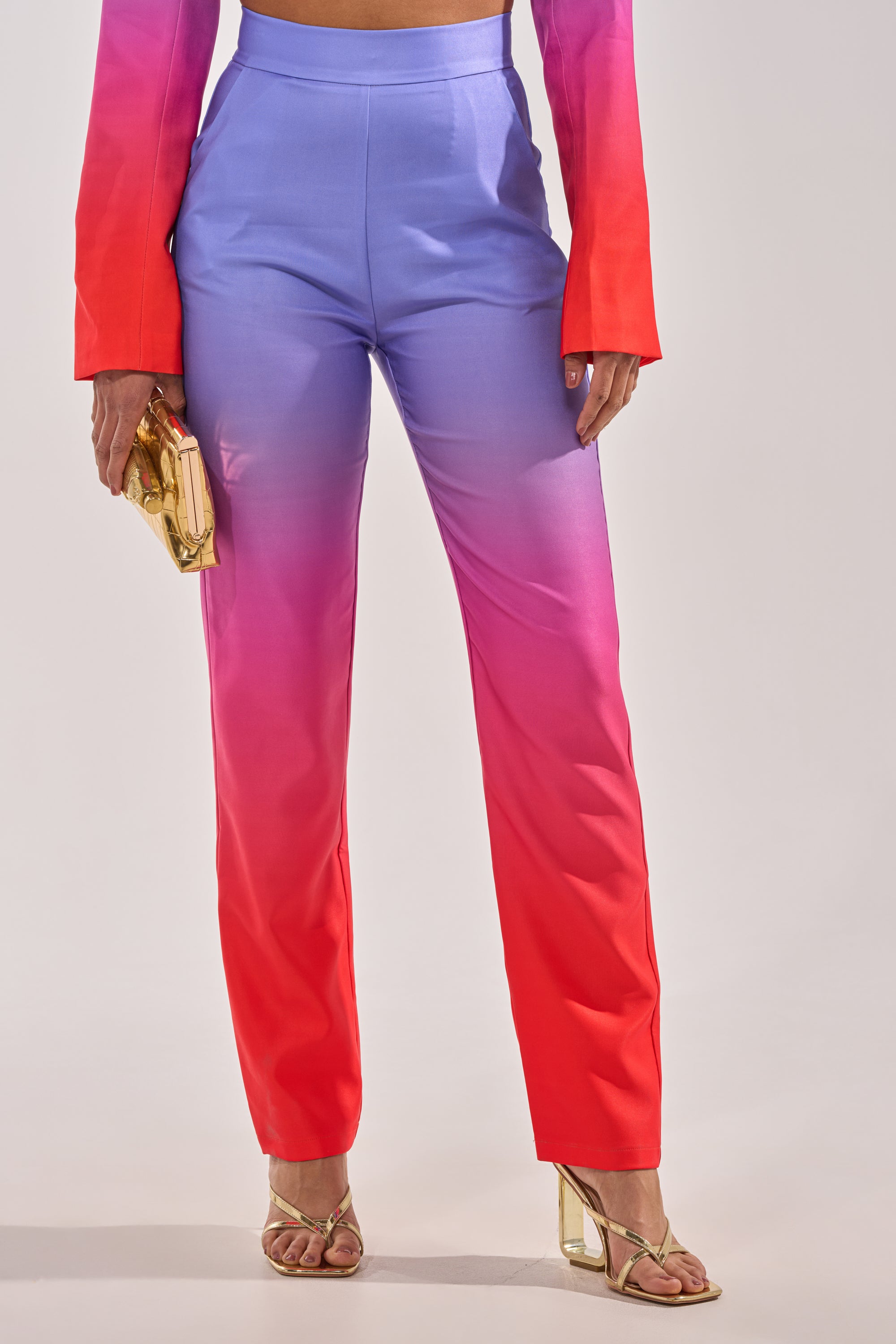 Someone in the SUNSET IN PARADISE OMBRE PANT, featuring a high-rise waist and a pink-to-purple gradient, paired with a matching long-sleeve top, gold heeled sandals, and a gold clutch, stands against a plain background. 1