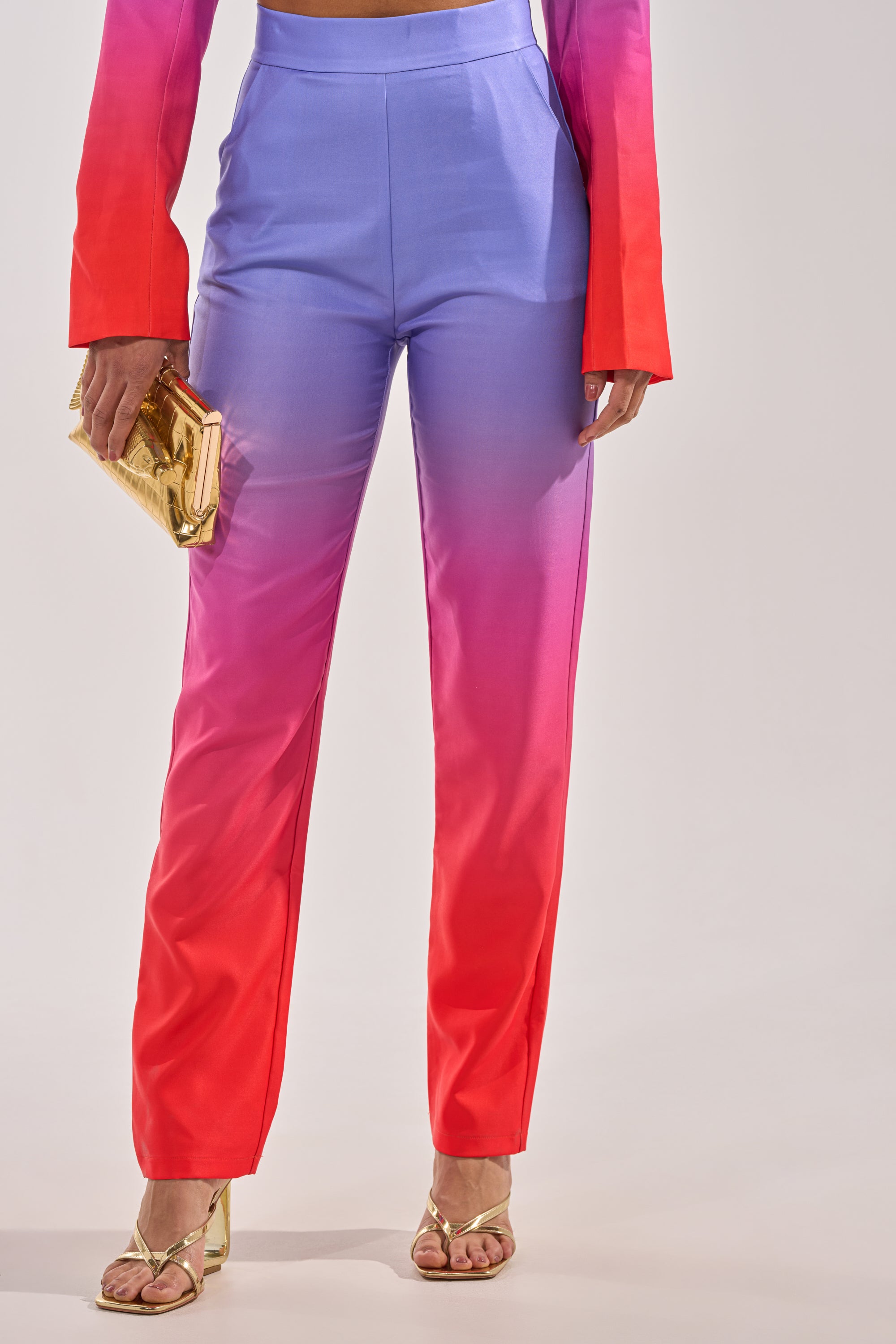 Wearing the SUNSET IN PARADISE OMBRE PANT, which fades from purple to pink and red, paired with a matching long-sleeve top. The look is styled with a gold clutch and strappy gold heels. 2