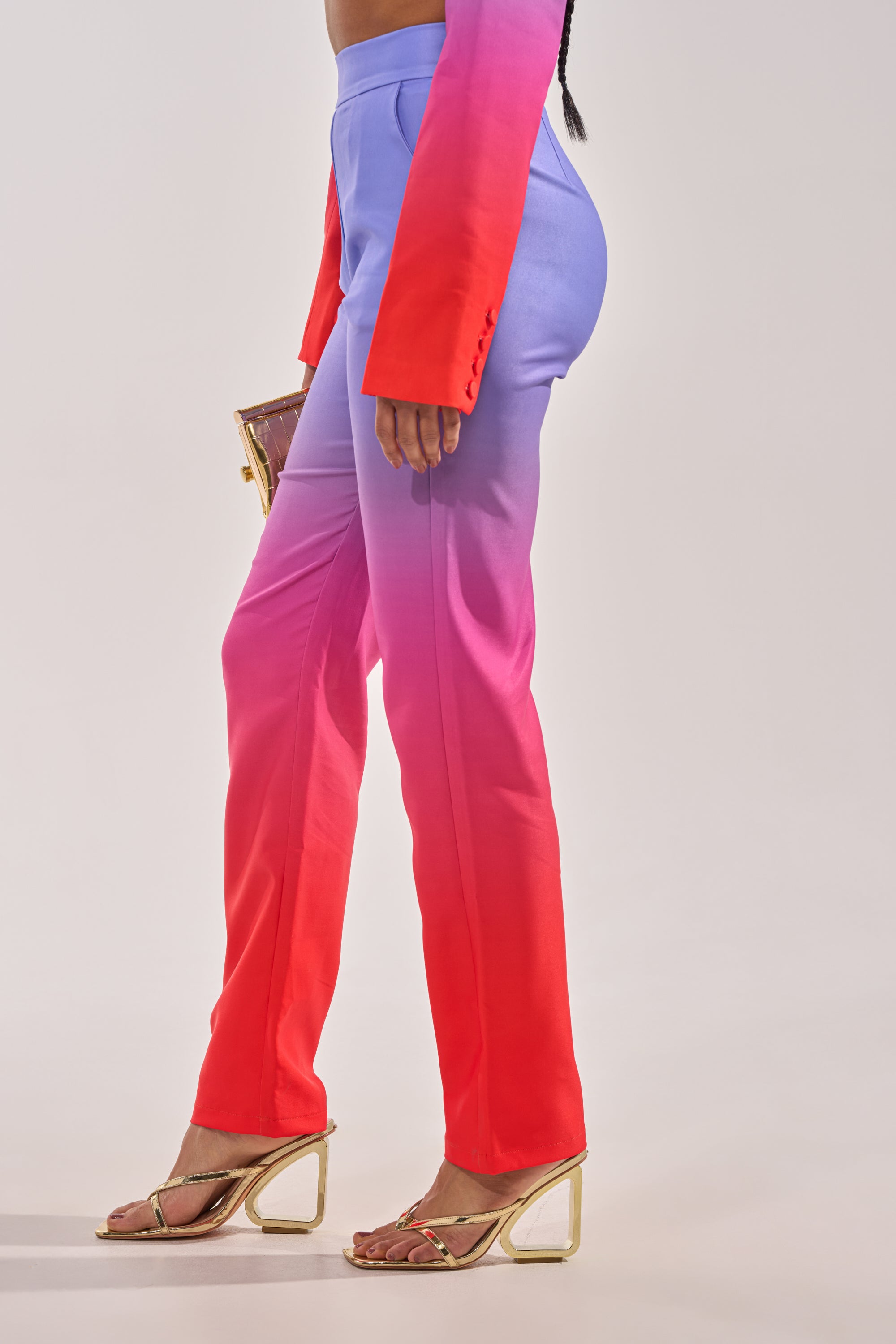Wearing the SUNSET IN PARADISE OMBRE PANT with a high-rise, straight-leg fit, plus a matching blazer, gold-heeled sandals, and a small clutch, someone poses against a light backdrop. 3