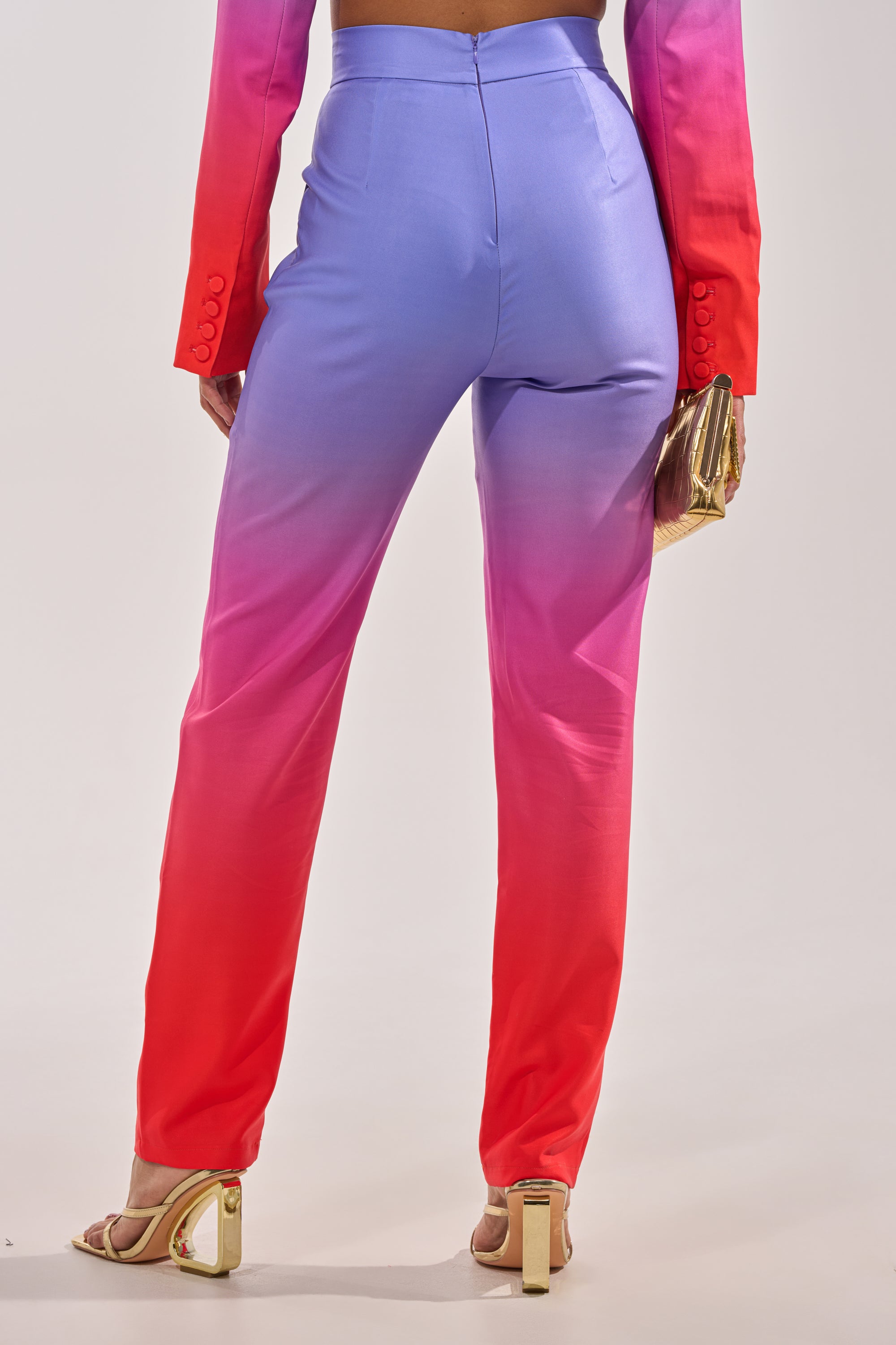 A person wears the SUNSET IN PARADISE OMBRE PANT with a matching red and purple blazer, gold heels, and a small gold clutch. The pants fade from light purple to red, and the image shows the outfit from the back. 4