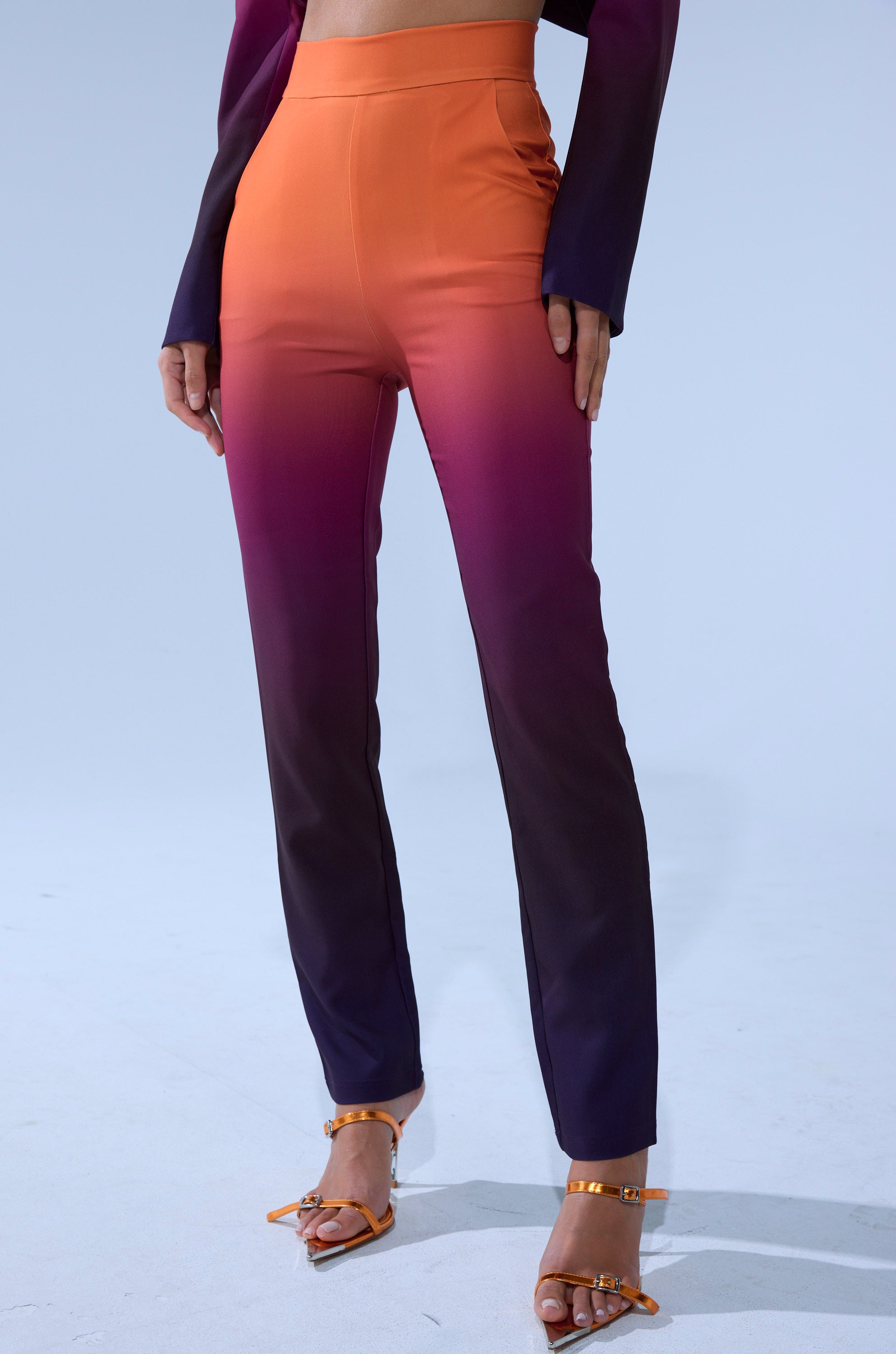 A person wears the RISE AND SHINE TROUSER, a high-rise pant with a gradient fade from orange to purple and black at the hem, styled with heeled sandals and a coordinating long-sleeve top.