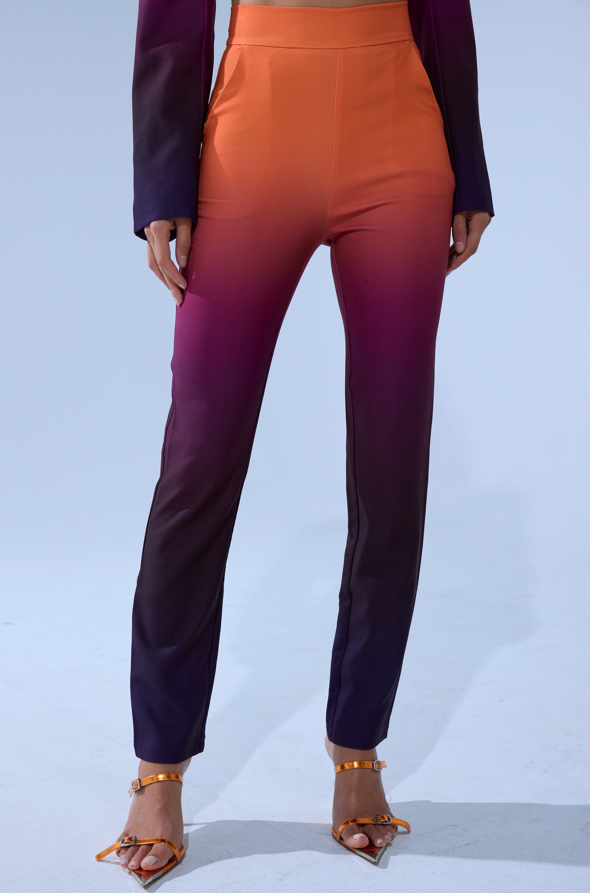 A person models the RISE AND SHINE TROUSER—high-rise pants with a gradient fade from orange at the waist to dark purple at the ankles—styled with strappy heels. The image is cropped to show from waist to feet.