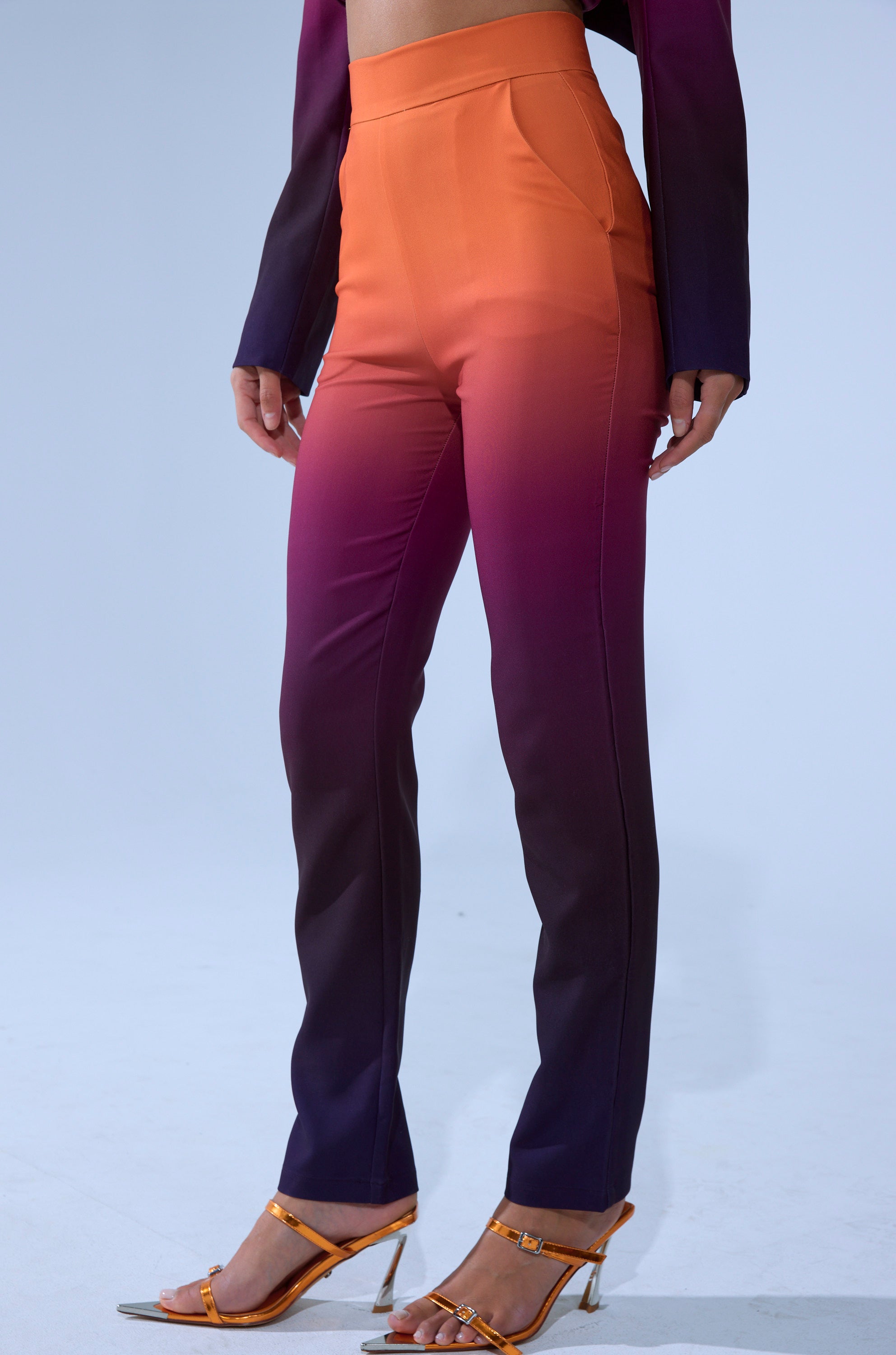 Someone wears the RISE AND SHINE TROUSER, a high-rise ombre style fading from orange at the waist to deep purple at the ankles, paired with orange heels and a coordinating long-sleeved top.